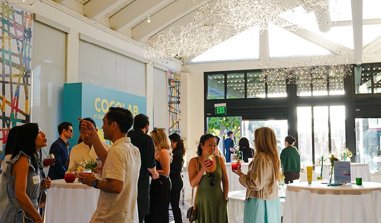 Guests standing and mingling at a cocktail reception in a modern event space with high-top tables and large windows.