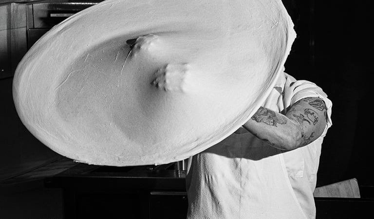 Black-and-white photo of a chef stretching a large sheet of pizza dough in a commercial kitchen.