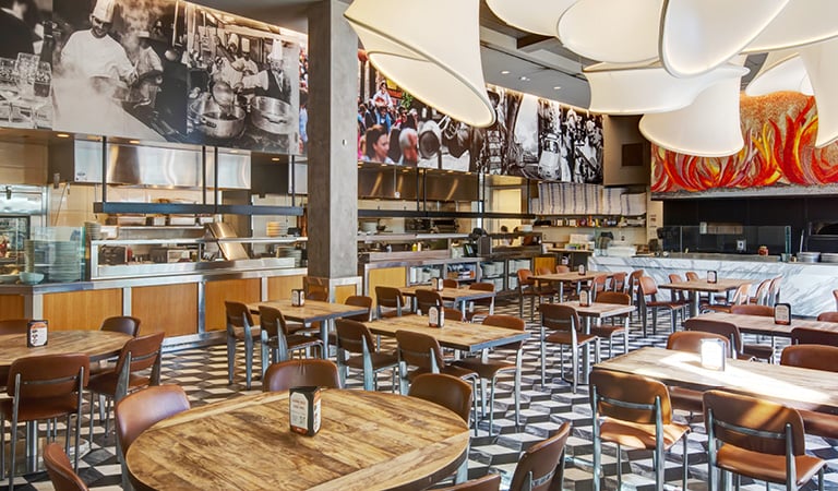 Spacious restaurant dining room with an open kitchen, wood tables, and pizza ovens beneath a colorful flame-themed mural.