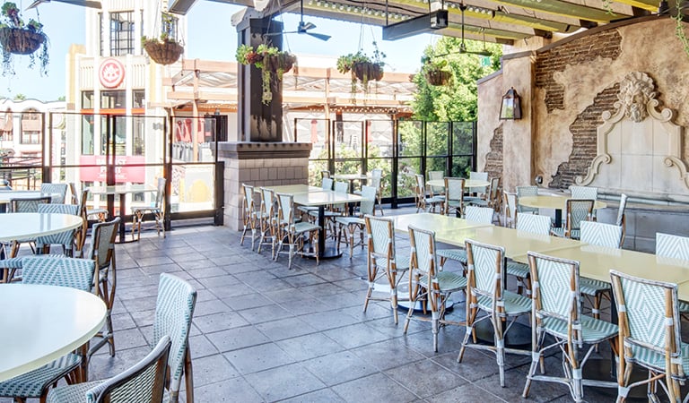 Bright covered patio dining area with café tables, hanging plants, and a decorative wall fountain overlooking an outdoor plaza.